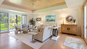 A spacious living room with a large window, a ceiling fan, a sofa set, a coffee table, a side table, a lamp, a painting, a round mirror, and a wooden cabinet.
