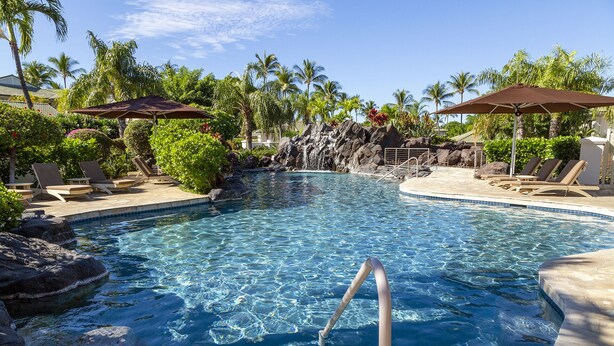 A clear pool with a rocky waterfall feature, surrounded by lounge chairs and umbrellas.