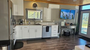 A modern kitchen with white cabinets, a built-in dishwasher, a coffee maker, and a large window. - Modern container cabin on 13 acre country estate (Sequim)