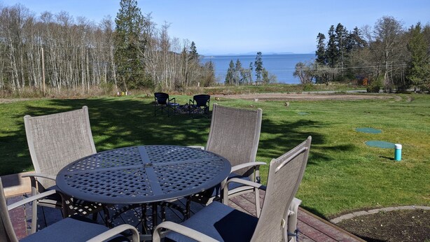 A round metal table with four chairs, a grassy lawn, and a view of the ocean. - Modern container cabin on 13 acre country estate (Sequim)