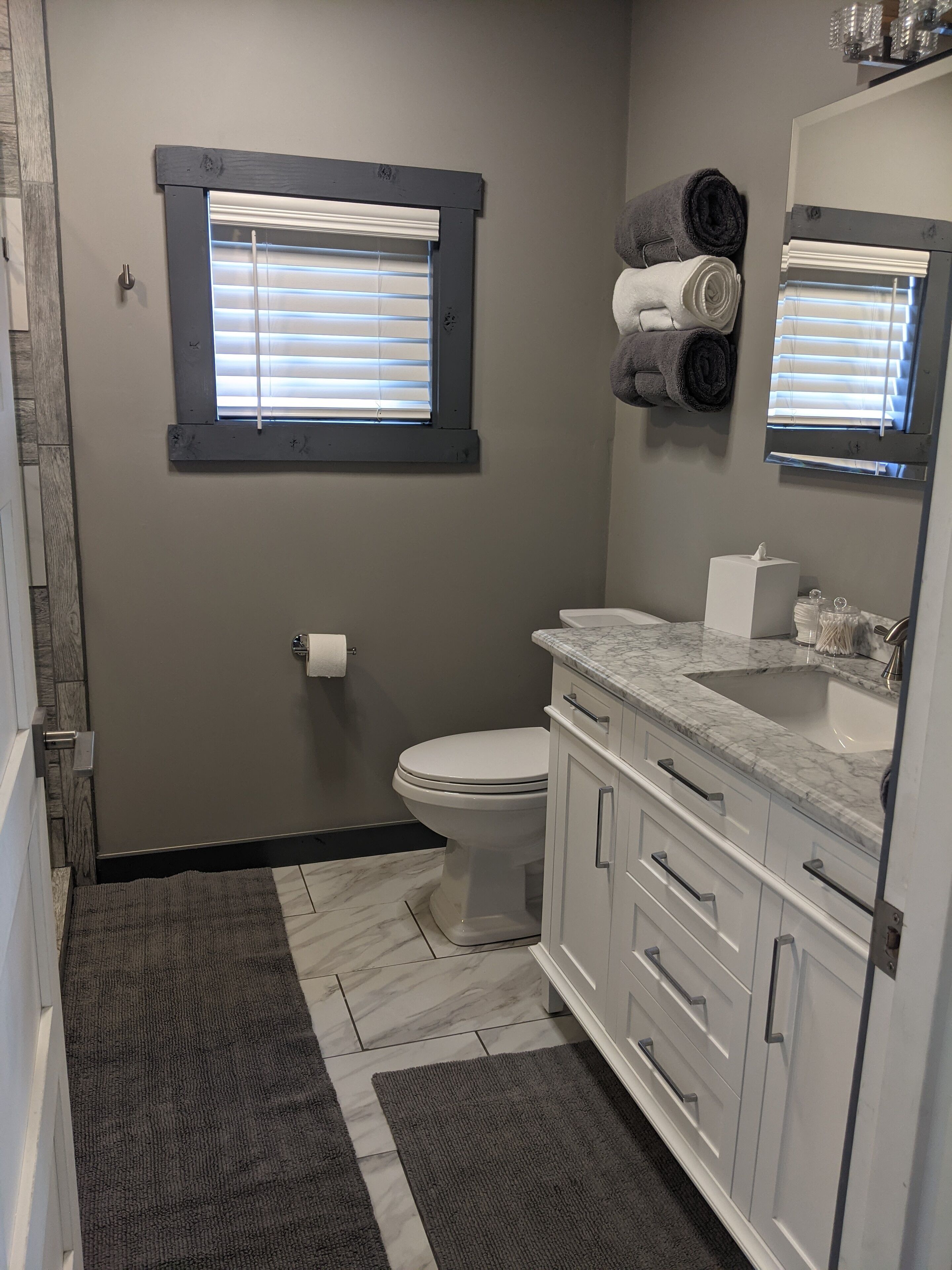 A bathroom with a toilet, a sink, a mirror, and a window with blinds.