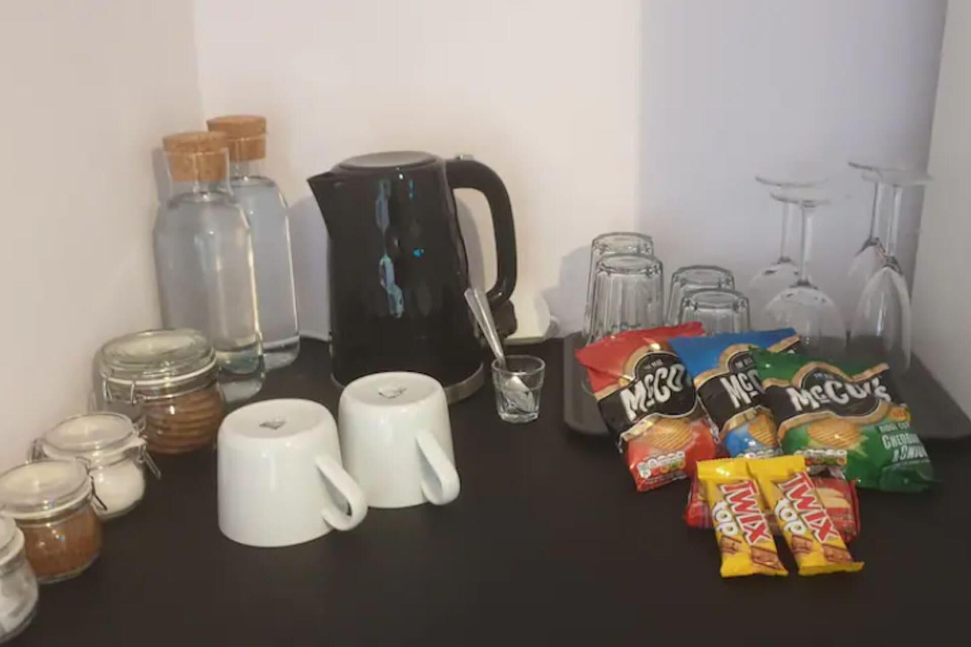 A kitchen counter with various items including jars, mugs, a coffee maker, glasses, and snack packages.
