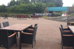 An outdoor seating area with wicker tables and chairs, a covered boat, and a stone wall in the background.