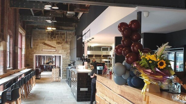Ein geräumiges Restaurant mit hohen Decken und freiliegenden Backsteinwänden. Eine lange Theke mit Hockern und ein Barbereich sind vorhanden. Rechts ist eine große Ballondekoration zu sehen.