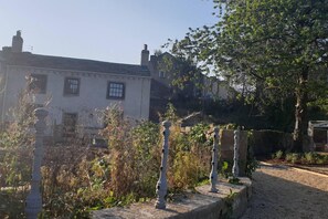 A building with a white facade and multiple chimneys, surrounded by overgrown vegetation and a gravel path.