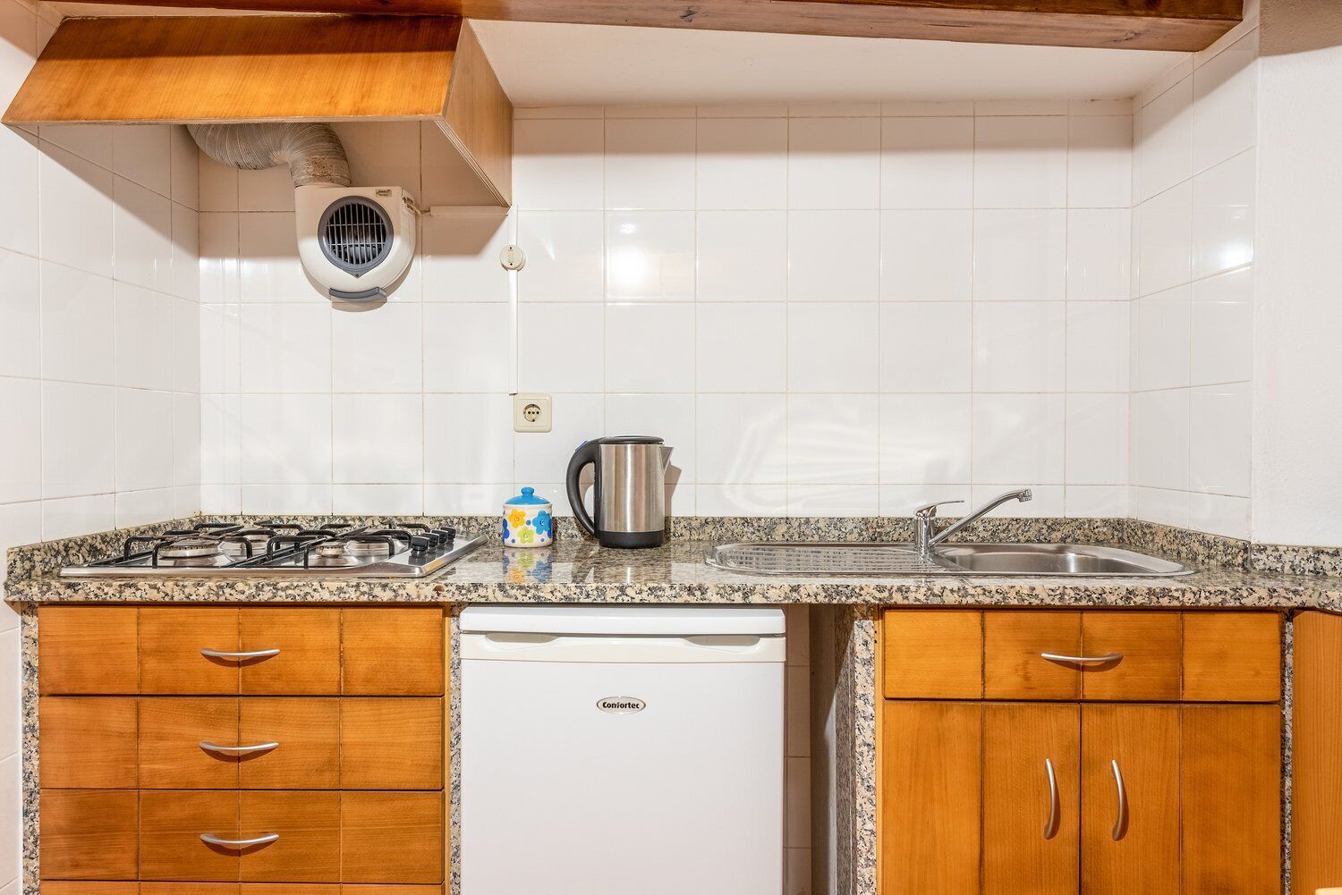 Una cuina amb parets revestides de rajoles blanques, taulell de granat, armaris de fusta, fogó a gas, cafetera i rentaplats.