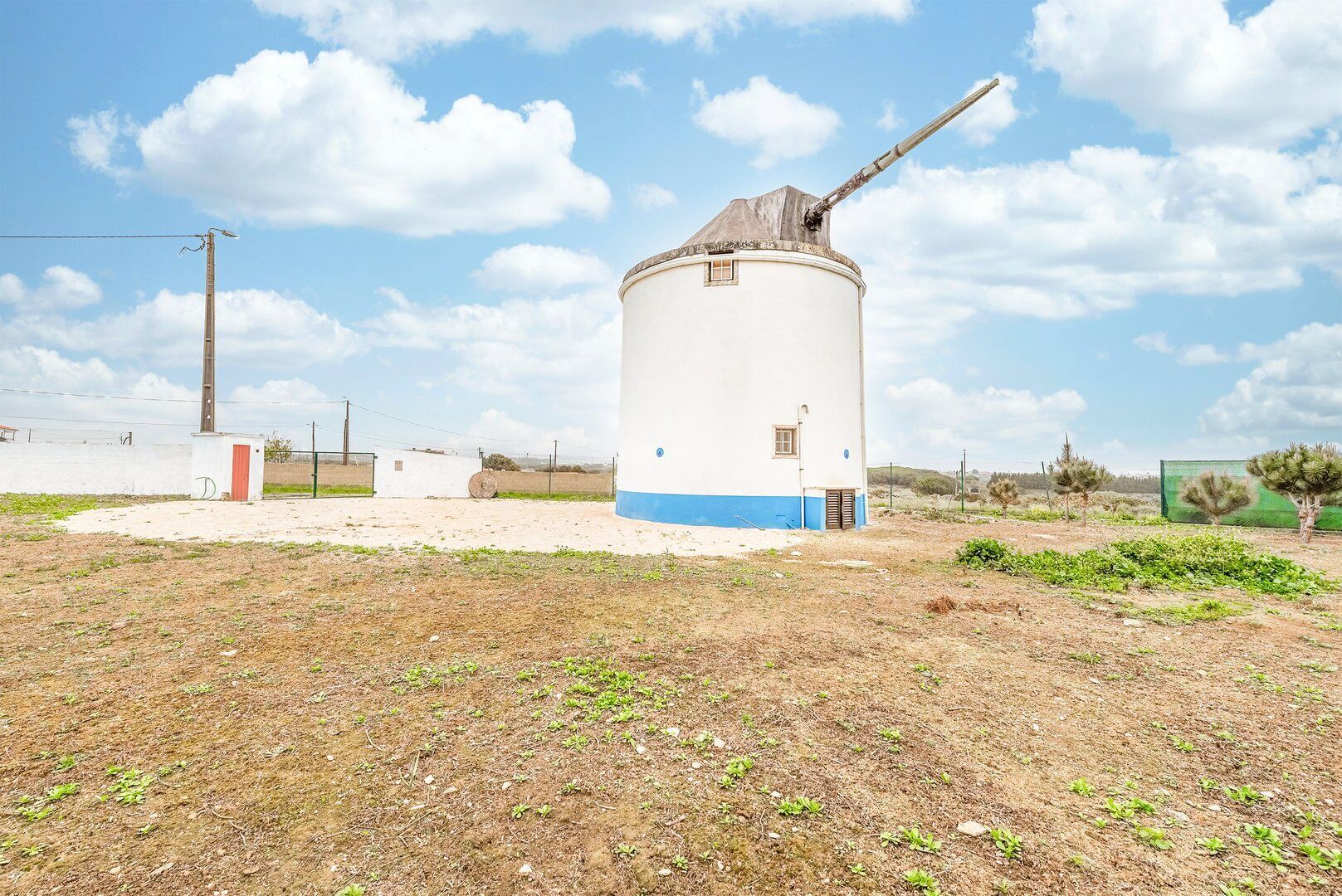 Eine Windmühle mit einem langen, horizontalen Arm, umgeben von einem eingezäunten Bereich mit spärlicher Vegetation und blauem Himmel.