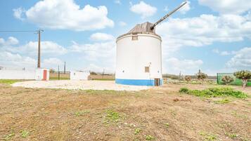 Eine Windmühle mit einem langen, horizontalen Arm, umgeben von einem eingezäunten Bereich mit spärlicher Vegetation und blauem Himmel.