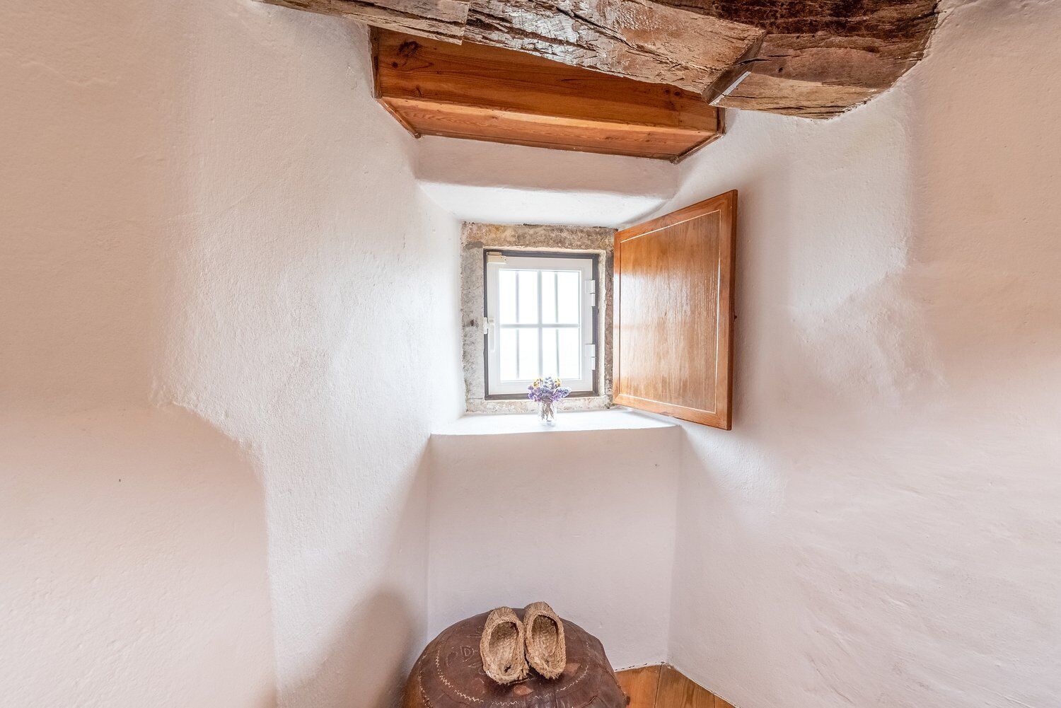 A narrow room with a wooden ceiling, a small window with a wooden door, and a pair of shoes on the floor.