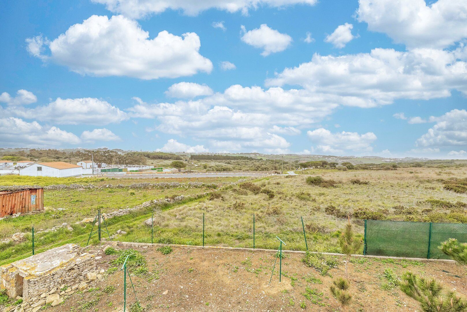 Paisaje rural con una cerca, un muro de piedra y algunos edificios a lo lejos.