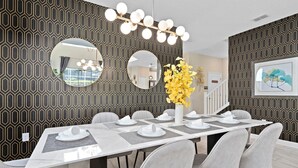 Un espace repas moderne avec une longue table dressée pour six personnes, entourée de chaises grises. Deux miroirs ronds sont accrochés au mur, ainsi qu’un arrangement floral jaune décoratif et une œuvre d’art encadrée au-dessus de la table.
