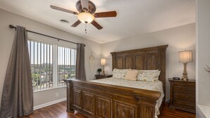 Ein Schlafzimmer mit einem großen Bett, einem Holz-Kopfteil, einem Deckenventilator, einem Nachttisch, einer Kommode und einem Fenster mit Vorhängen.