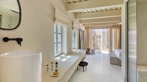 A modern bathroom with a large white sink, a mirror, and a window. There is a bench and a small table with toiletries. - Azade Chania (Chania)