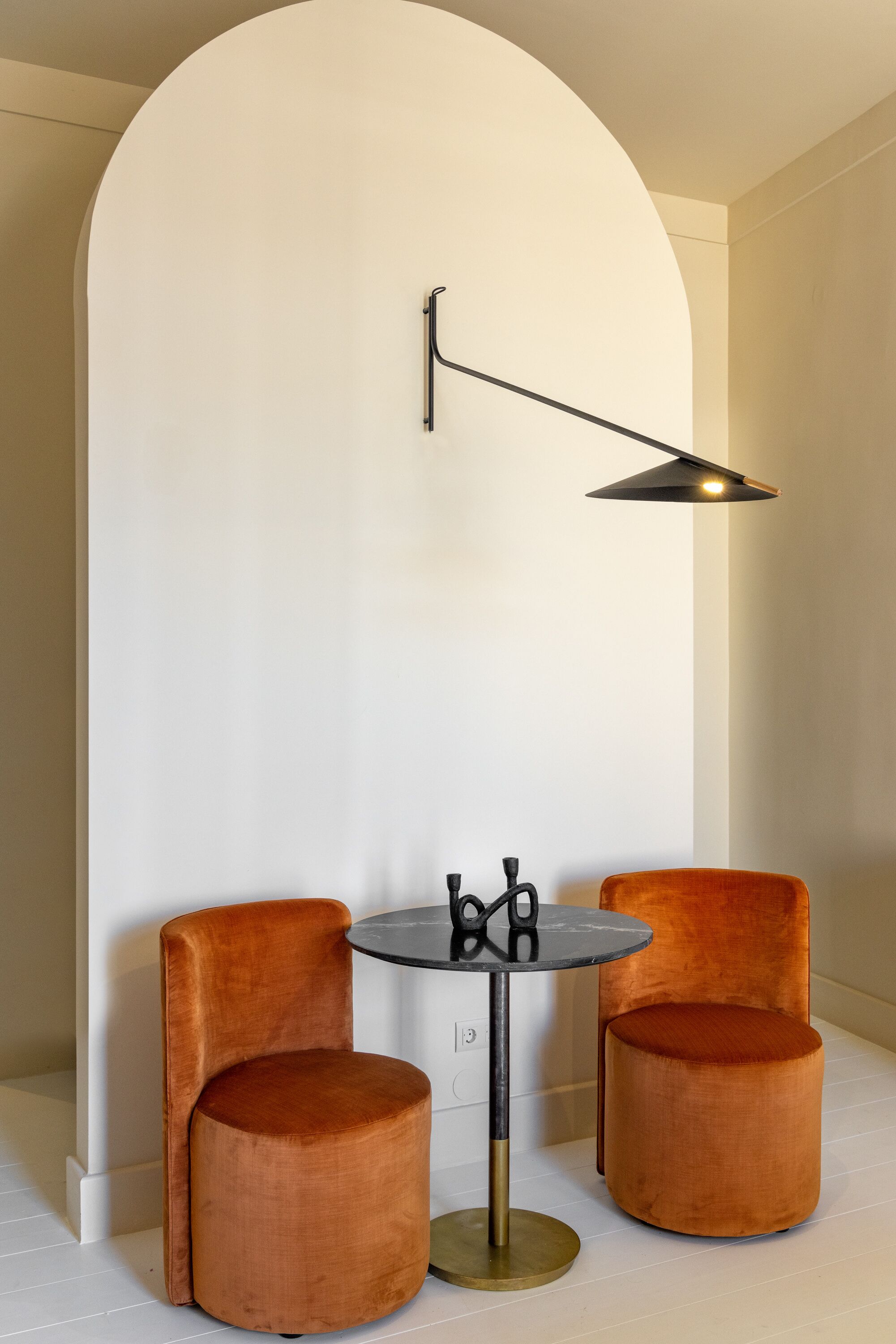 A modern room with a white wall, a round table with a black lamp, and two orange cushioned chairs.
