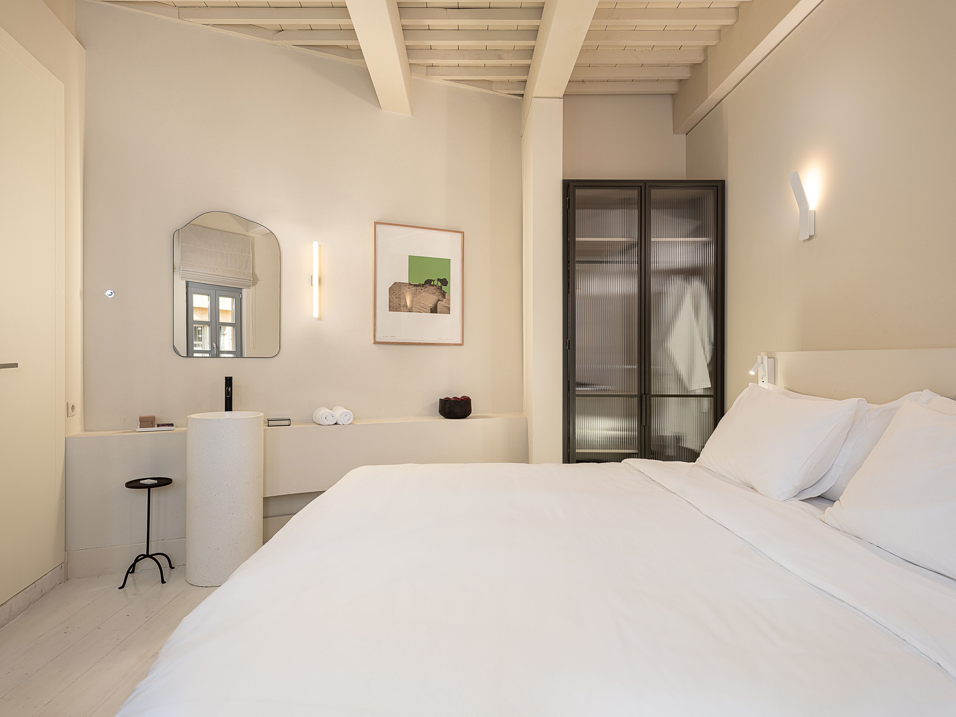 A modern hotel room with a large bed, a small bedside table, a mirror, a framed picture on the wall, and a sliding door leading to a bathroom.