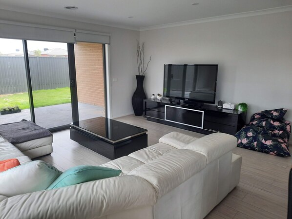 A modern living room with a large TV, a sofa, and a sliding glass door leading to a patio. - The Jewel of Seachange | Modern Family Retreat Minutes from the Beach (Saint Leonards)