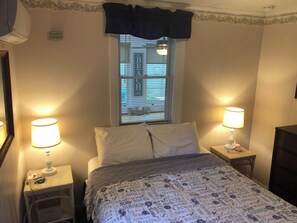 A bedroom with a bed, two bedside tables, a window with blinds, and a wall-mounted air conditioner.
