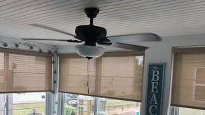 A cozy room with a ceiling fan, a table, chairs, a green storage unit, and a view of the outdoors through large windows with blinds. - 4 Nights Left -Perfect Family-Friendly Condo 5 Houses from the Ocean-Beach Block (Dewey Beach)