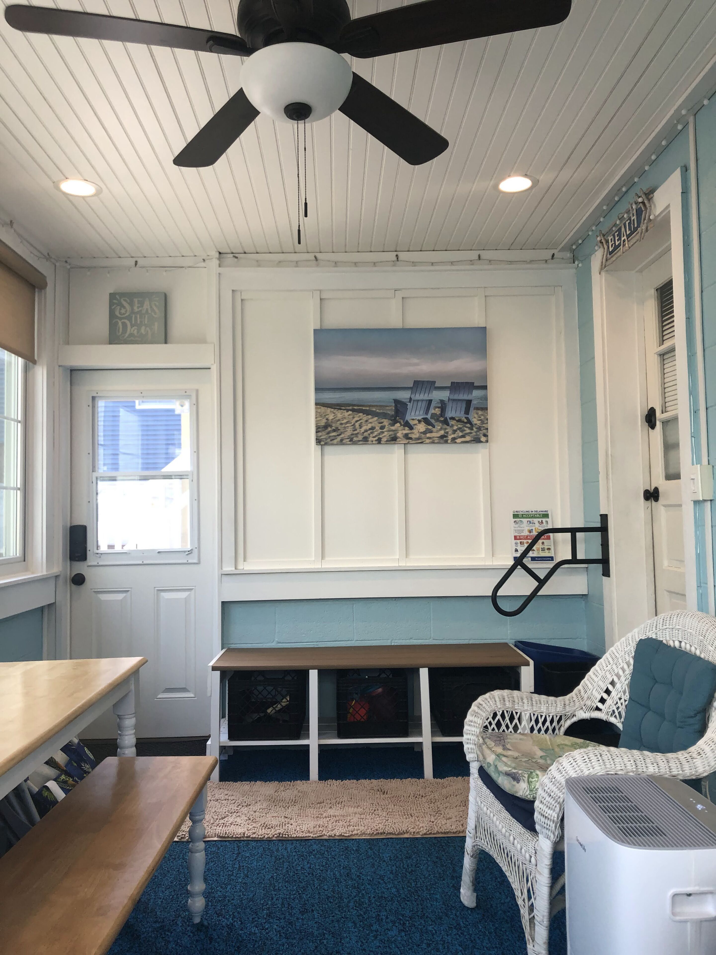 A room with a ceiling fan, a painting of a beach scene, a white wicker chair, a wooden table, and a blue carpet.