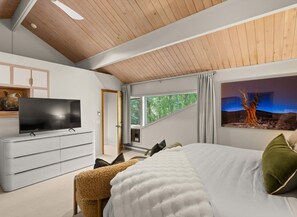 A bedroom with a bed, a TV on a white dresser, a window with a view of trees, and a painting on the wall.