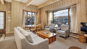 Una sala de estar acogedora con paredes de madera y ventanas amplias, un sofá, sillones, una mesa de centro y un televisor.