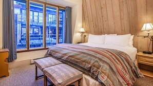 Habitación de hotel con una cama grande, una banca, dos mesitas de noche con lámparas y vistas a la ciudad a través de amplios ventanales.