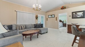 A spacious living room with a grey sectional sofa, a round coffee table, and a chandelier. There is a dining area with a table and chairs, and a kitchen visible in the background.