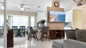 A modern living room with a grey sofa, a flat-screen TV mounted on the wall, a dining area with a table and chairs, and a kitchen area with a marble countertop.