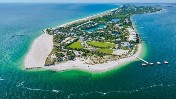 An aerial view of a coastal resort with a long beachfront, clear blue waters, and a well-organized layout of buildings and green spaces.