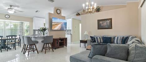Un salón amplio con un sofá gris, una mesa de comedor con sillas, un televisor montado en la pared y una araña.