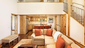 A spacious living area with a high ceiling, wooden accents, a sofa set, and a kitchen in the background. - Fifth Avenue Unit 202 | 2 Bedroom + Loft Condo, Huge Windows & Views. 2 Blocks to Skiing! (Aspen)