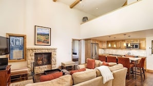A spacious living room with a fireplace, a dining area with bar stools, and a kitchen with wooden cabinets and modern appliances. - Fifth Avenue Unit 202 | 2 Bedroom + Loft Condo, Huge Windows & Views. 2 Blocks to Skiing! (Aspen)