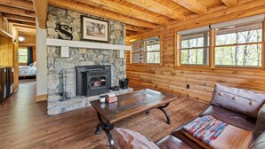A log cabin interior with a stone fireplace, wooden ceiling beams, and large windows.