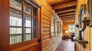 A log cabin interior with a wooden door, large windows, a small table with a lamp, and a fireplace.