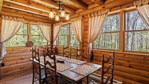 A wooden cabin with a dining table set for six, large windows with curtains, and a wooden ceiling with exposed beams.