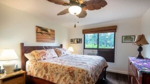 Un dormitorio con una cama, una mesita de noche, un escritorio, un ventilador de techo, una ventana y un cuadro colgado en la pared.