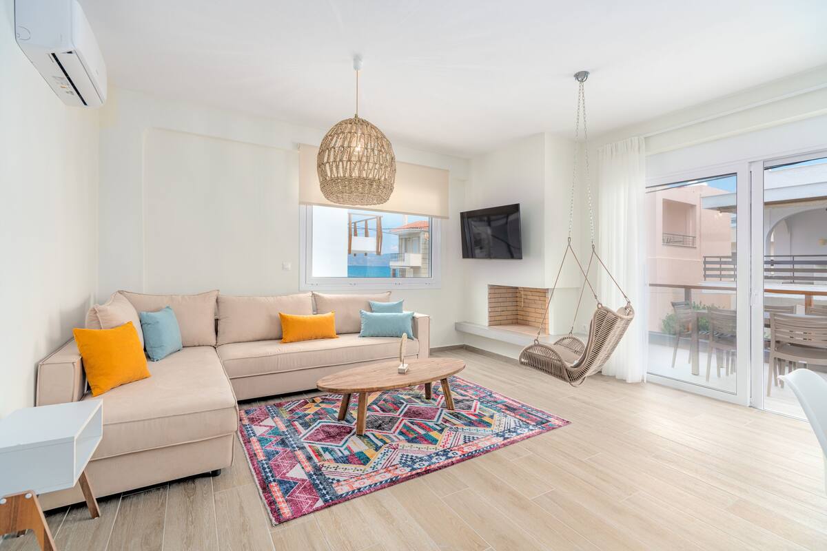 A modern living room with a beige sofa, colorful cushions, a patterned rug, a hanging wicker chair, and a large window offering a view of a building.