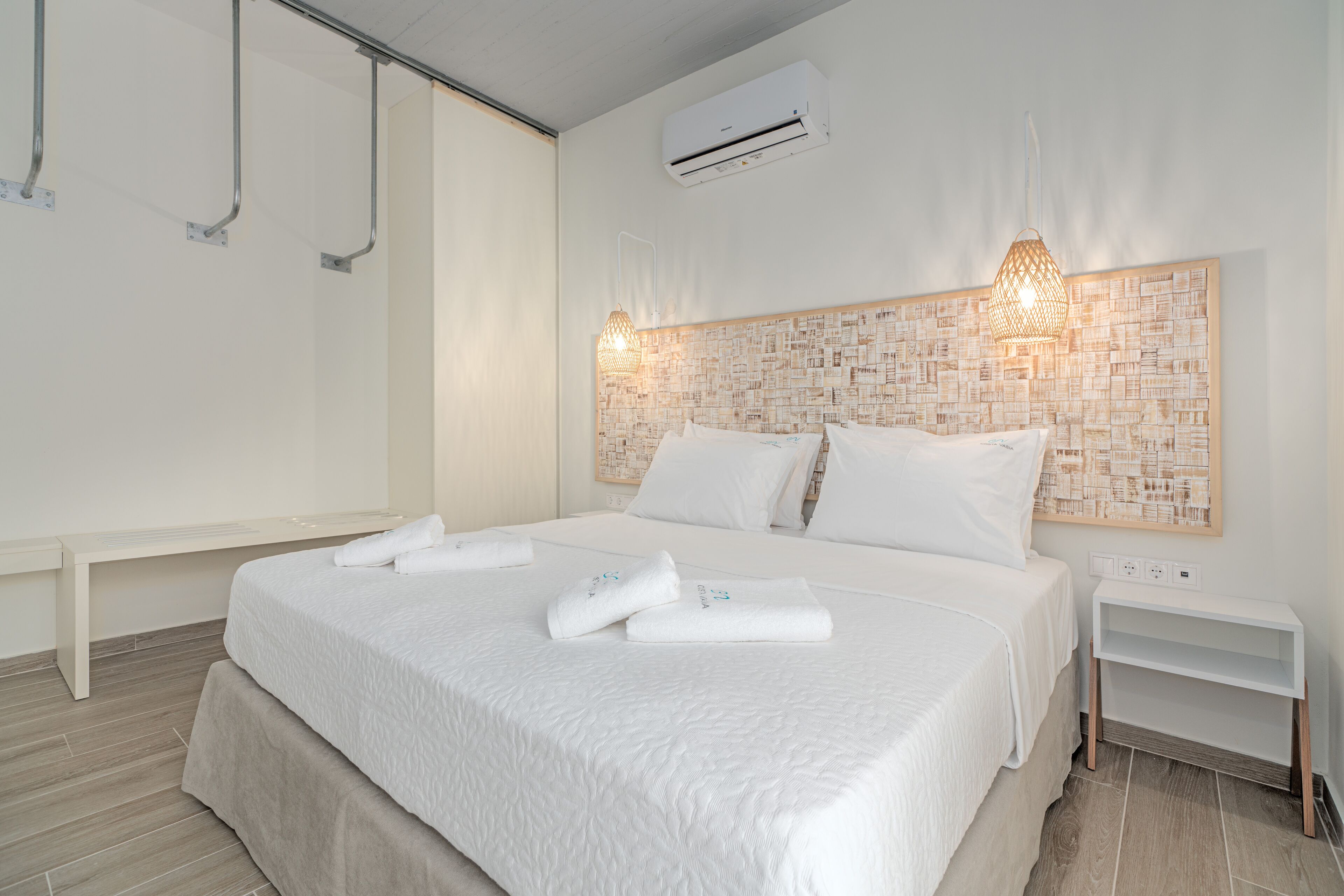 A neatly arranged hotel room with a large bed, white linens, a wall-mounted air conditioner, and a bedside table with a lamp.
