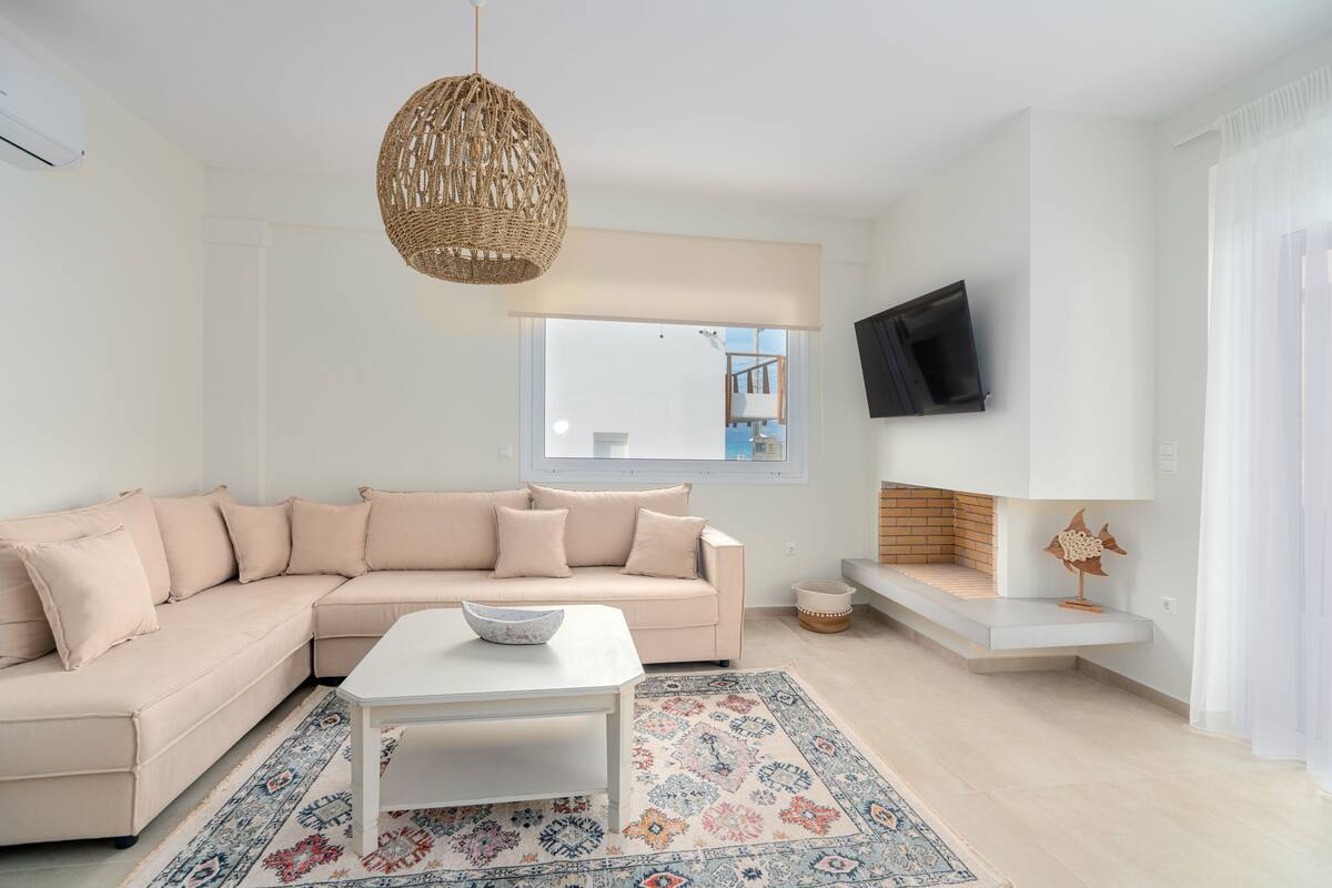 A modern living room with a beige sectional sofa, a white coffee table, a patterned rug, a wicker pendant light, a flat-screen TV mounted on the wall, and a small fireplace with a white mantel.