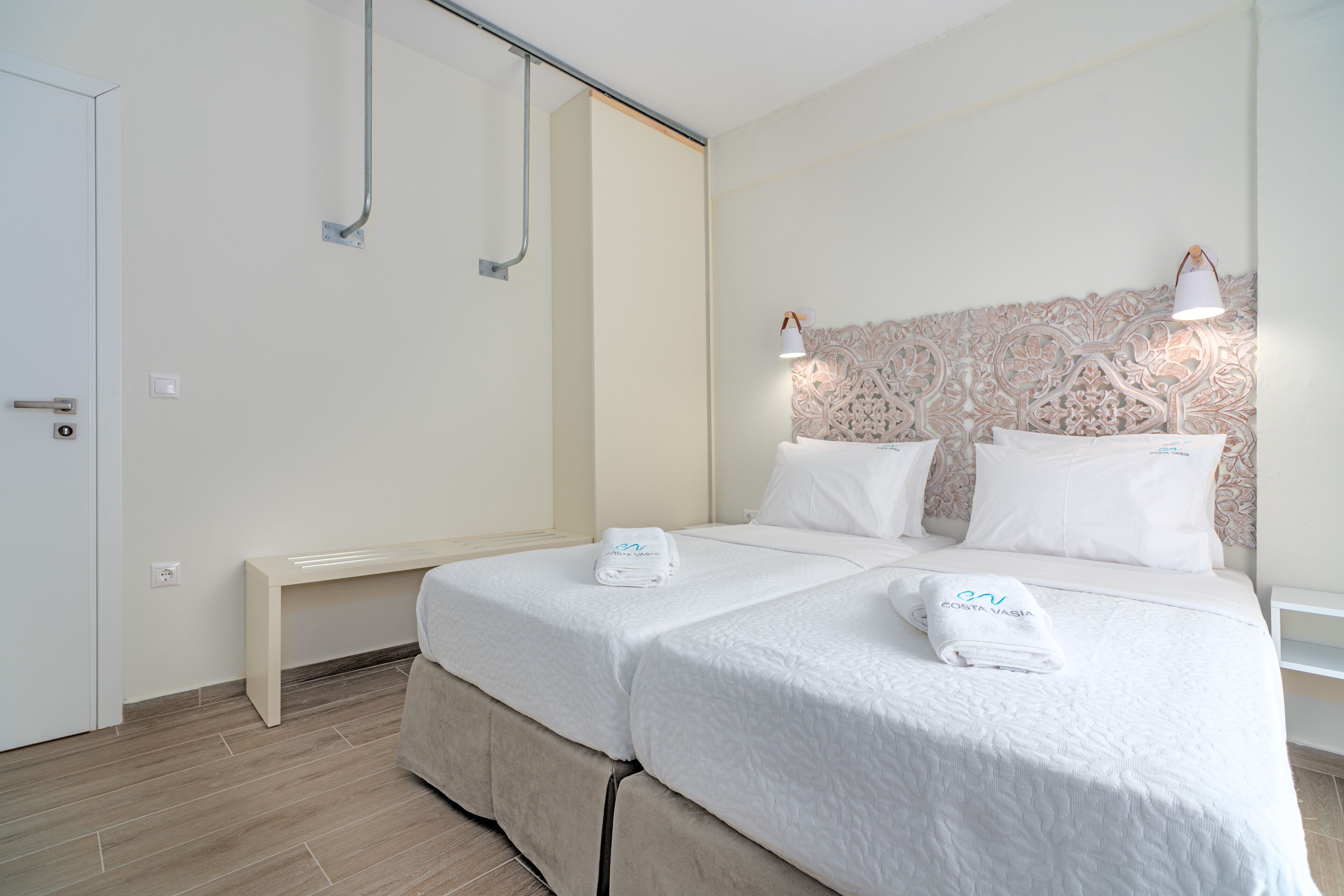 Una habitación de hotel con una cama, dos mesitas de noche, un banco y una lámpara fijada a la pared.