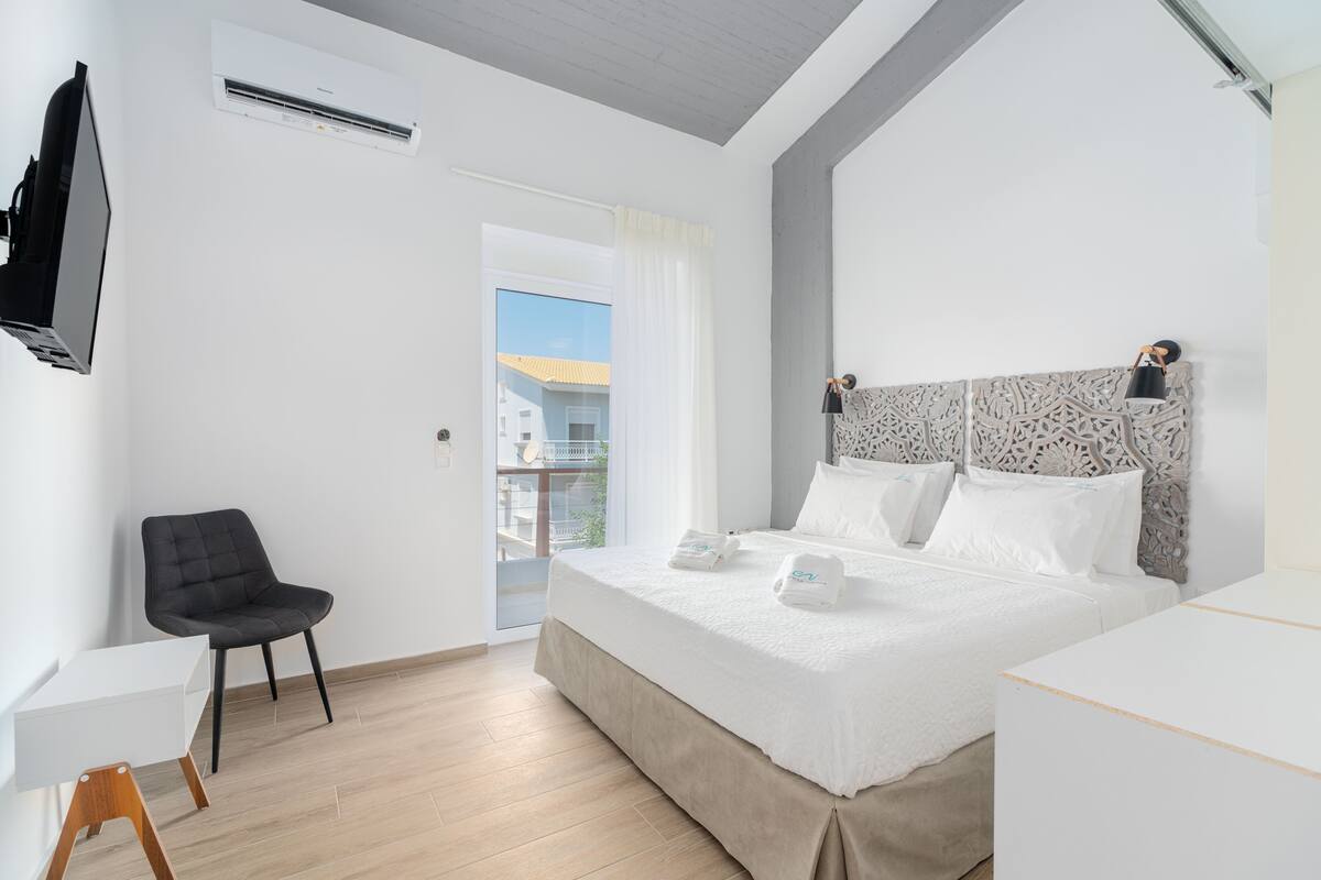 A modern hotel room with a large bed, a flat-screen TV, a chair, and a balcony with a view.
