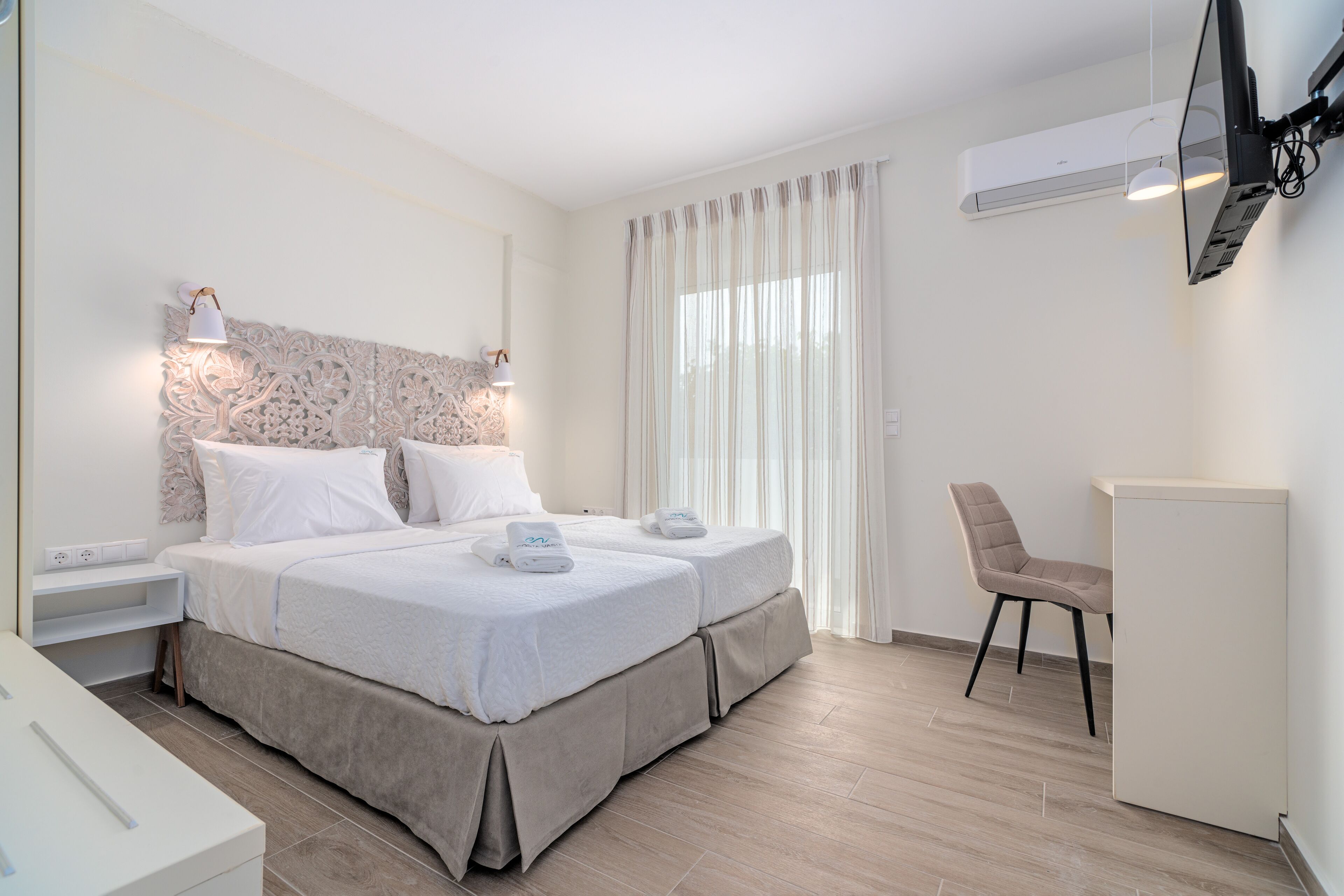 A hotel room with a bed, bedside tables, a chair, a desk, and an air conditioning unit.