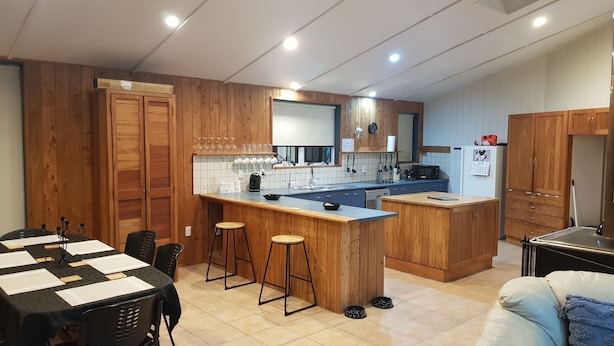 Una cocina moderna con armarios de madera, una isla central con taburetes, un comedor con mesa y sillas, y un refrigerador con un calendario.