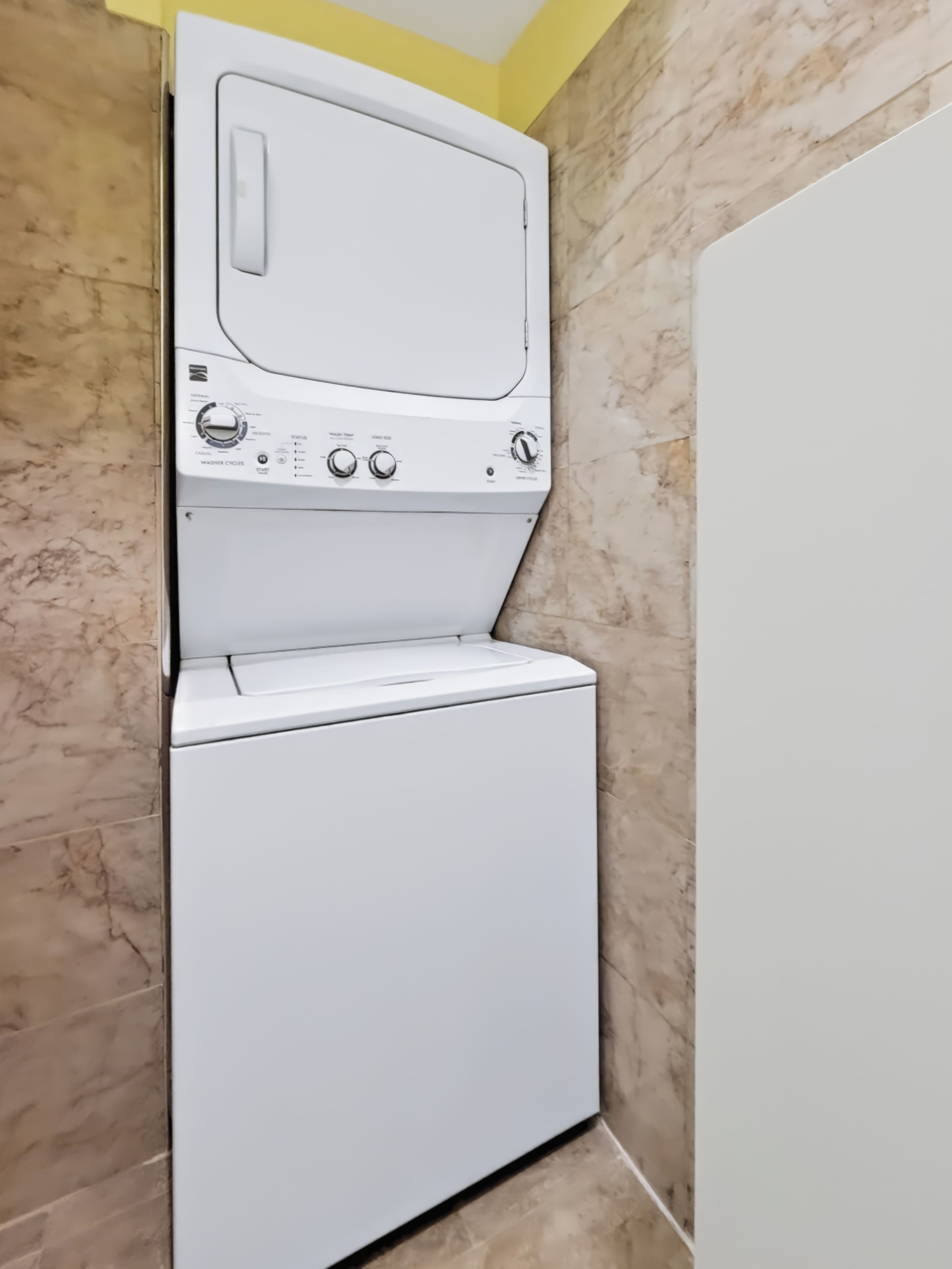 Een wasmachine en droger met een voorlaadklep, bedieningsknoppen en een klein display, voor een gele plafond en marmerachtige wandtegels.