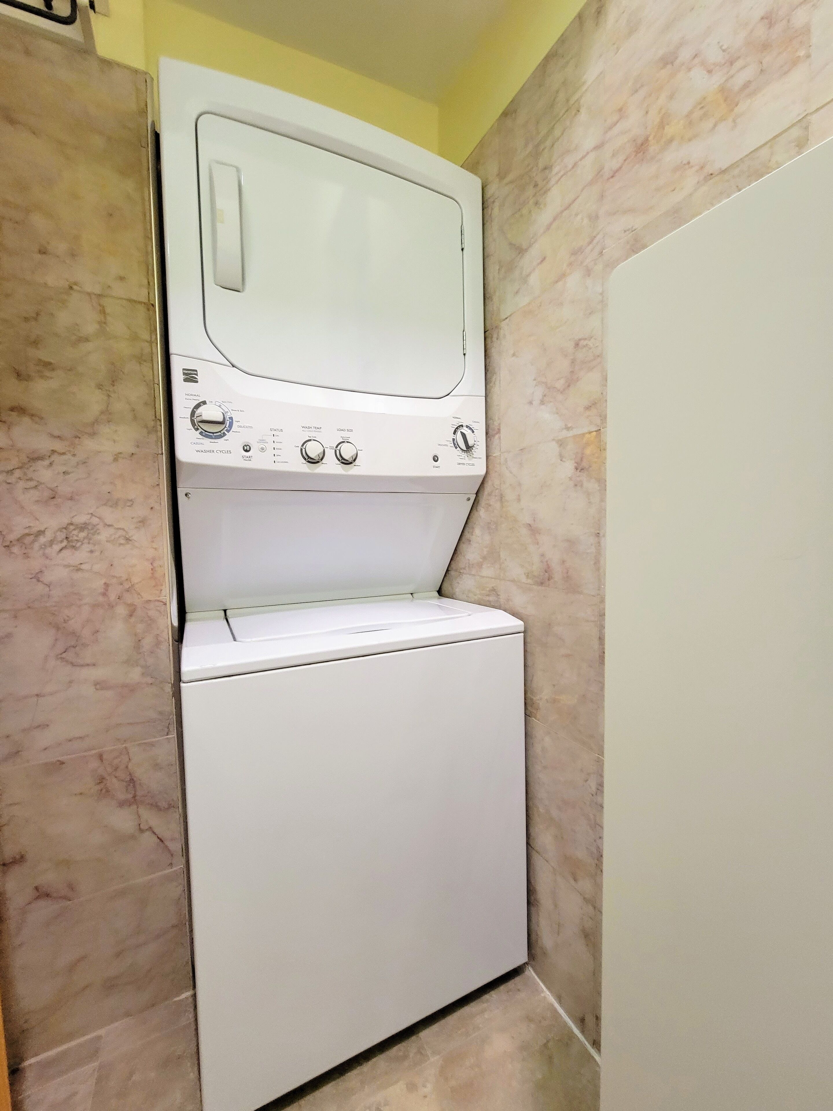 Une petite machine à laver blanche à chargement frontal, installée dans une petite buanderie aux murs en marbre.