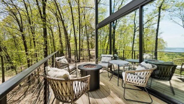 Eine Holzterrasse mit Korbmöbeln und einer Feuerstelle, umgeben von Glaswänden mit Blick auf einen Wald.