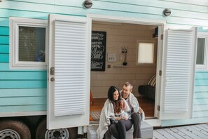 A couple sitting on a porch of a turquoise mobile home with open white shutters, a blackboard with motivational quotes, and a small table with a candle. - Lovely Tiny House Nestled in the Eucalyptus trees (San Luis Obispo)