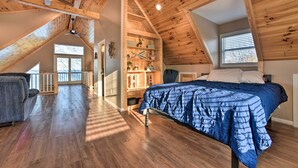 A loft bedroom with a sloped ceiling, wooden beams, a bed with a blue bedspread, a desk, and a chair. - Lakefront Lexington Escape: Steps to Beach! (Lexington)