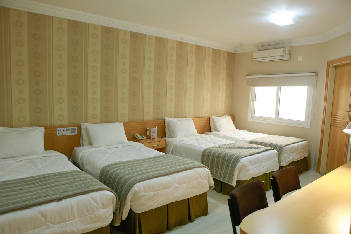 Habitación de hotel con tres camas individuales, cabecera de madera, mesita de noche con lámpara y ventana con persianas.