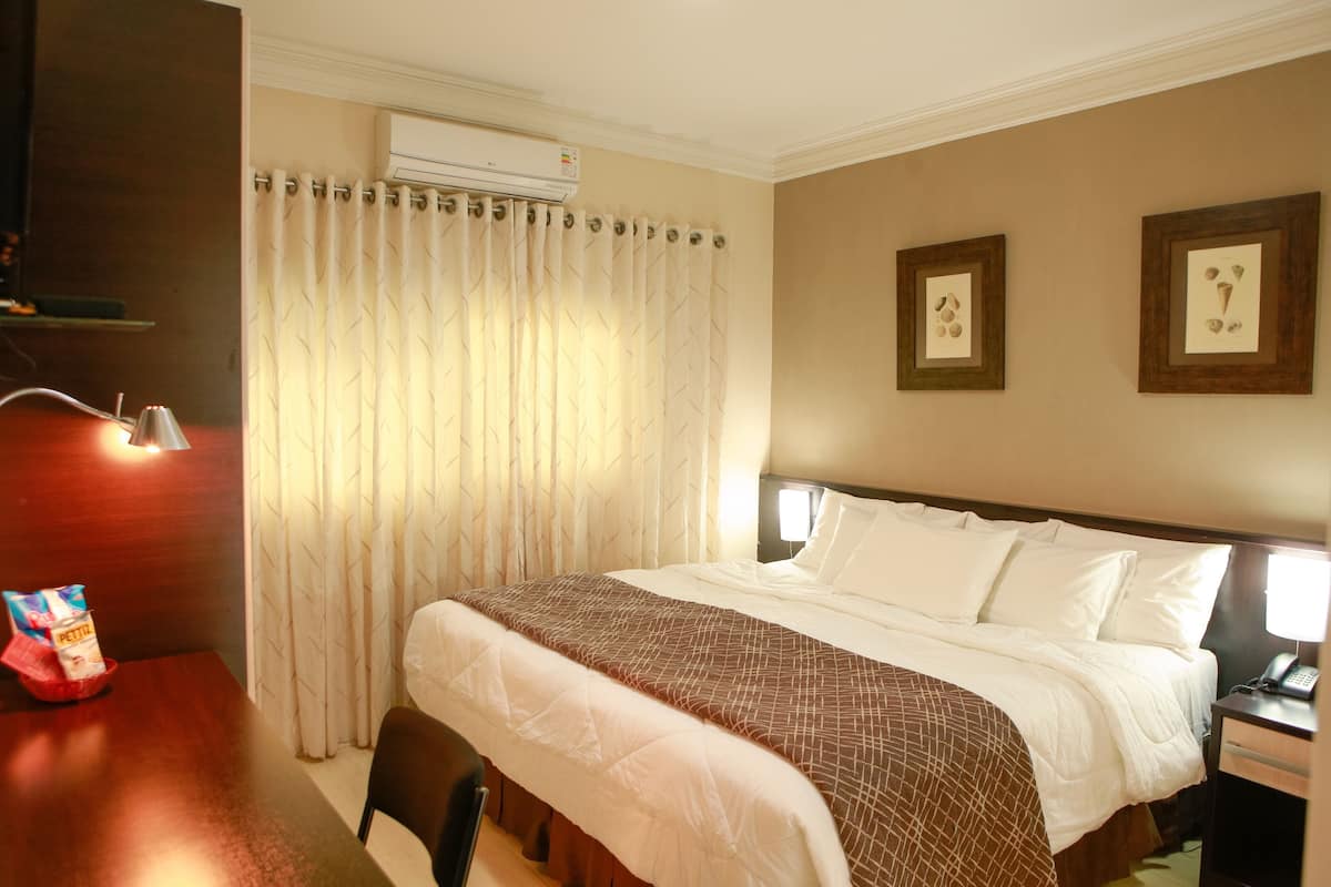 Habitación de hotel con una cama grande, una mesita de noche con lámpara, una silla y una pequeña mesa con aperitivos.